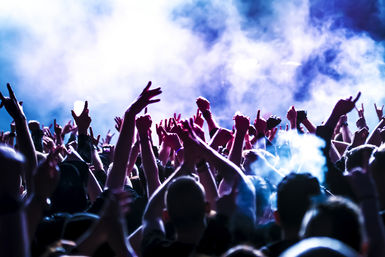 Energetic live music crowd with hands raised, glowing purple-blue stage lights and smoky atmosphere