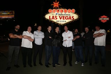Group of nine men standing with arms crossed beneath the illuminated 'Welcome to Fabulous Las Vegas Nevada' sign at night, neon-lit landmark in the background.