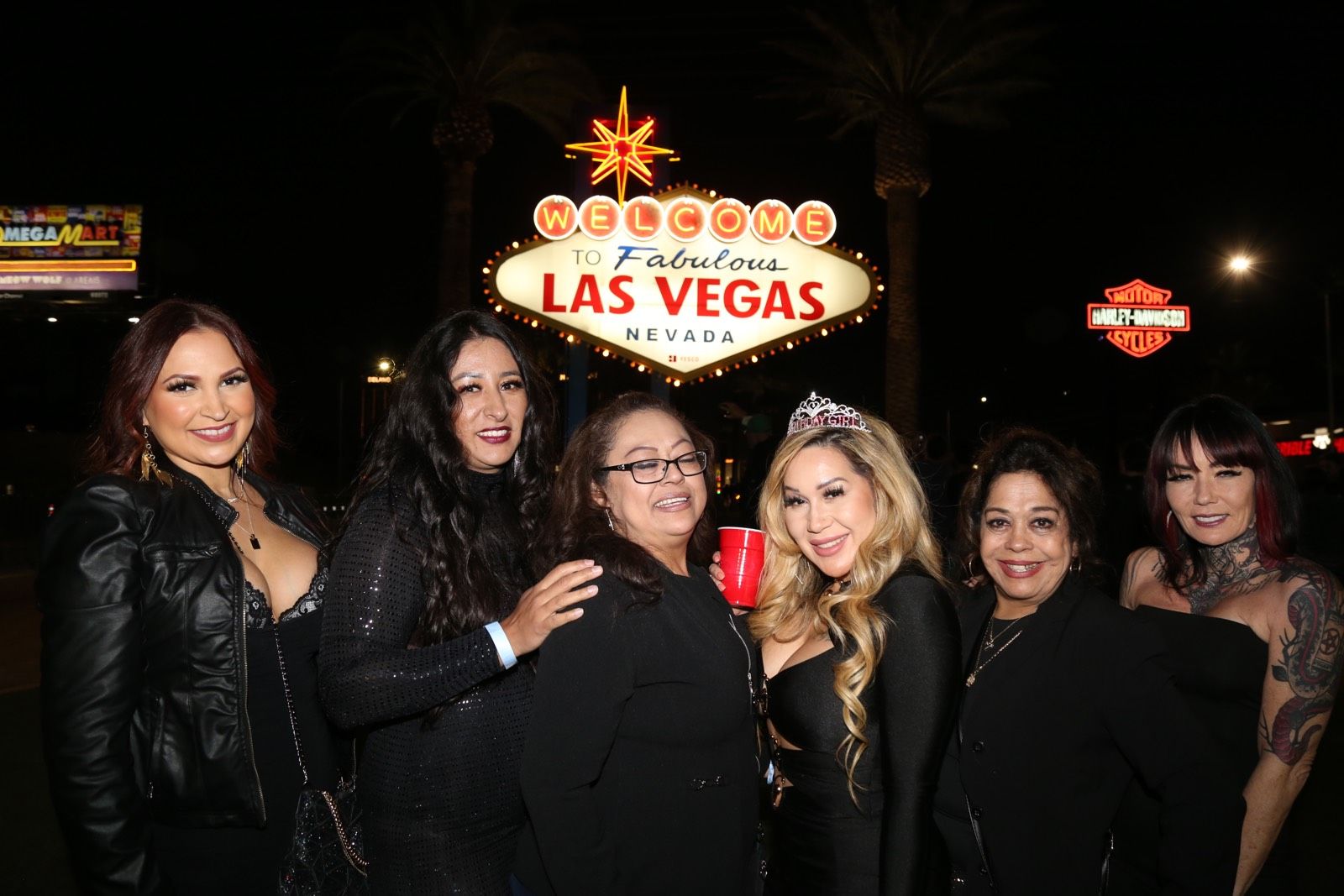 Group of six women in evening outfits smiling and posing at night in front of the illuminated Welcome to Fabulous Las Vegas, Nevada sign; one woman wears a tiara and holds a red cup with palm trees and neon lights in the background.