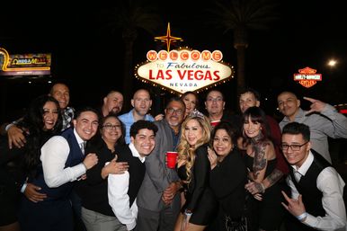 Smiling group of partygoers posing at night in front of the illuminated Welcome to Fabulous Las Vegas Nevada sign, some holding red cups and a woman wearing a tiara