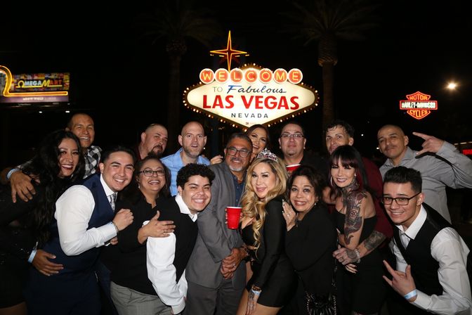 Smiling group of partygoers posing at night in front of the illuminated Welcome to Fabulous Las Vegas Nevada sign, some holding red cups and a woman wearing a tiara