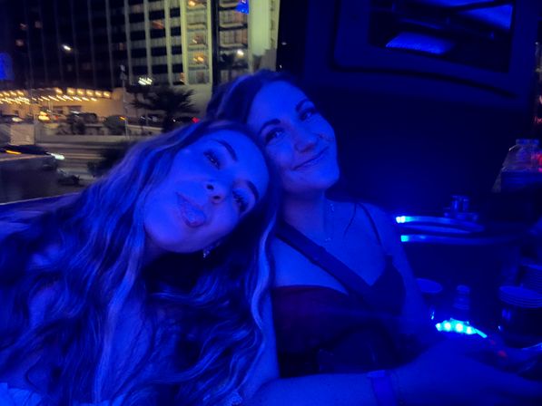 Two friends posing for a playful blue-lit selfie in a car at night, with city hotel windows and street lights glowing outside the window.