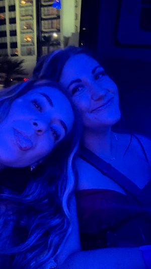 Two friends posing for a playful blue-lit selfie in a car at night, with city hotel windows and street lights glowing outside the window.