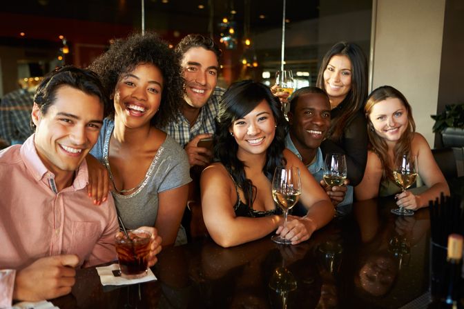 Diverse group of friends smiling at a bar, holding wine glasses and a cocktail during a lively night out in a cozy urban bar setting.