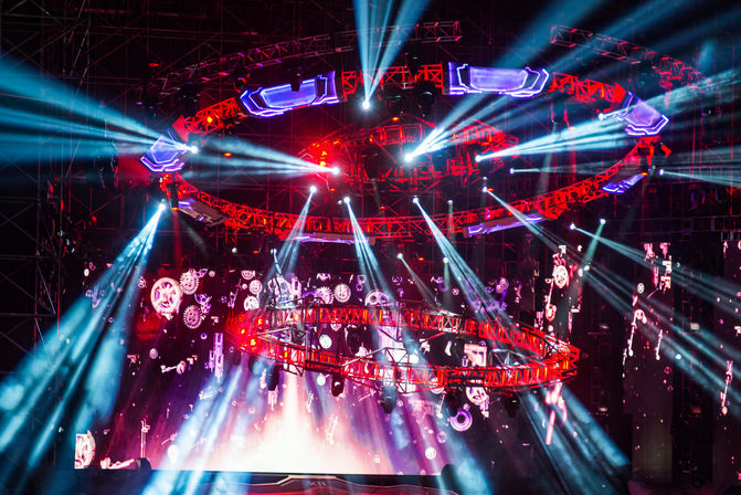 Vibrant concert stage at night with red circular trusses and blue-white spotlights radiating from a suspended lighting rig over an LED backdrop showing animated gears, evoking a high-energy music festival or live show.