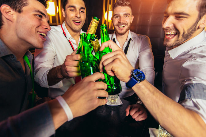 Group of friends in dress shirts smiling and clinking green beer bottles at a dimly lit bar on a night out