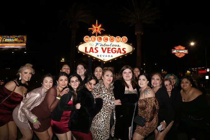 Group of women posing and smiling at night in front of the illuminated "Welcome to Fabulous Las Vegas Nevada" sign, dressed for a night out under palm trees and neon lights.