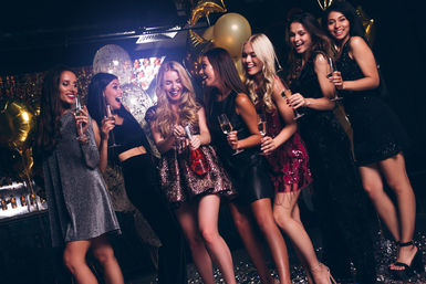 Seven women in festive party dresses cheering with champagne flutes and a popping bottle amid gold balloons and confetti at a lively nightclub celebration
