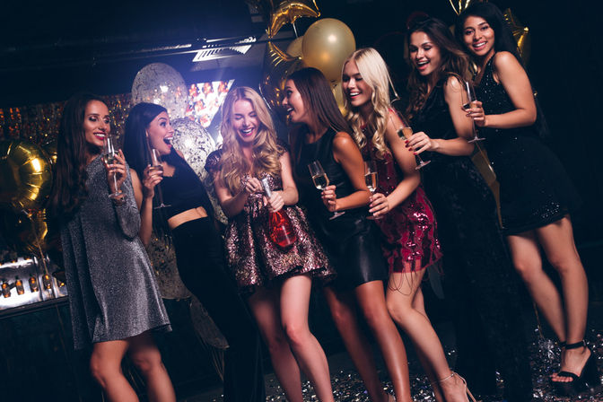 Seven women in festive party dresses cheering with champagne flutes and a popping bottle amid gold balloons and confetti at a lively nightclub celebration