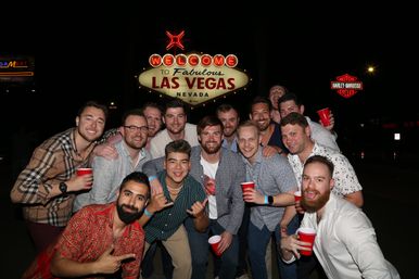 Smiling group of men holding red cups posing at night in front of the illuminated Welcome to Fabulous Las Vegas sign