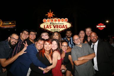 Large group of smiling friends posing and hugging at night in front of the illuminated "Welcome to Fabulous Las Vegas Nevada" sign on the Las Vegas Strip.