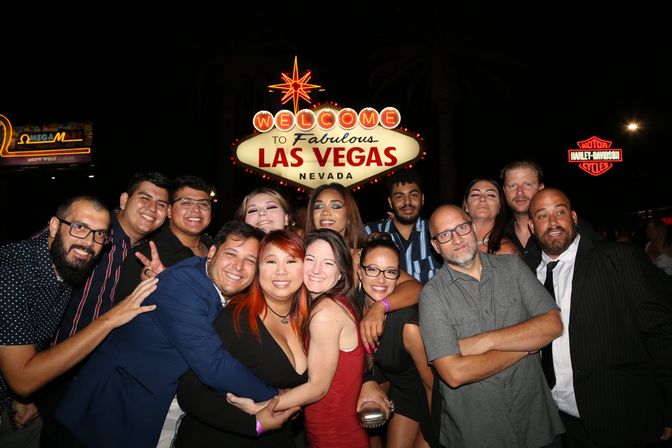 Large group of smiling friends posing and hugging at night in front of the illuminated "Welcome to Fabulous Las Vegas Nevada" sign on the Las Vegas Strip.