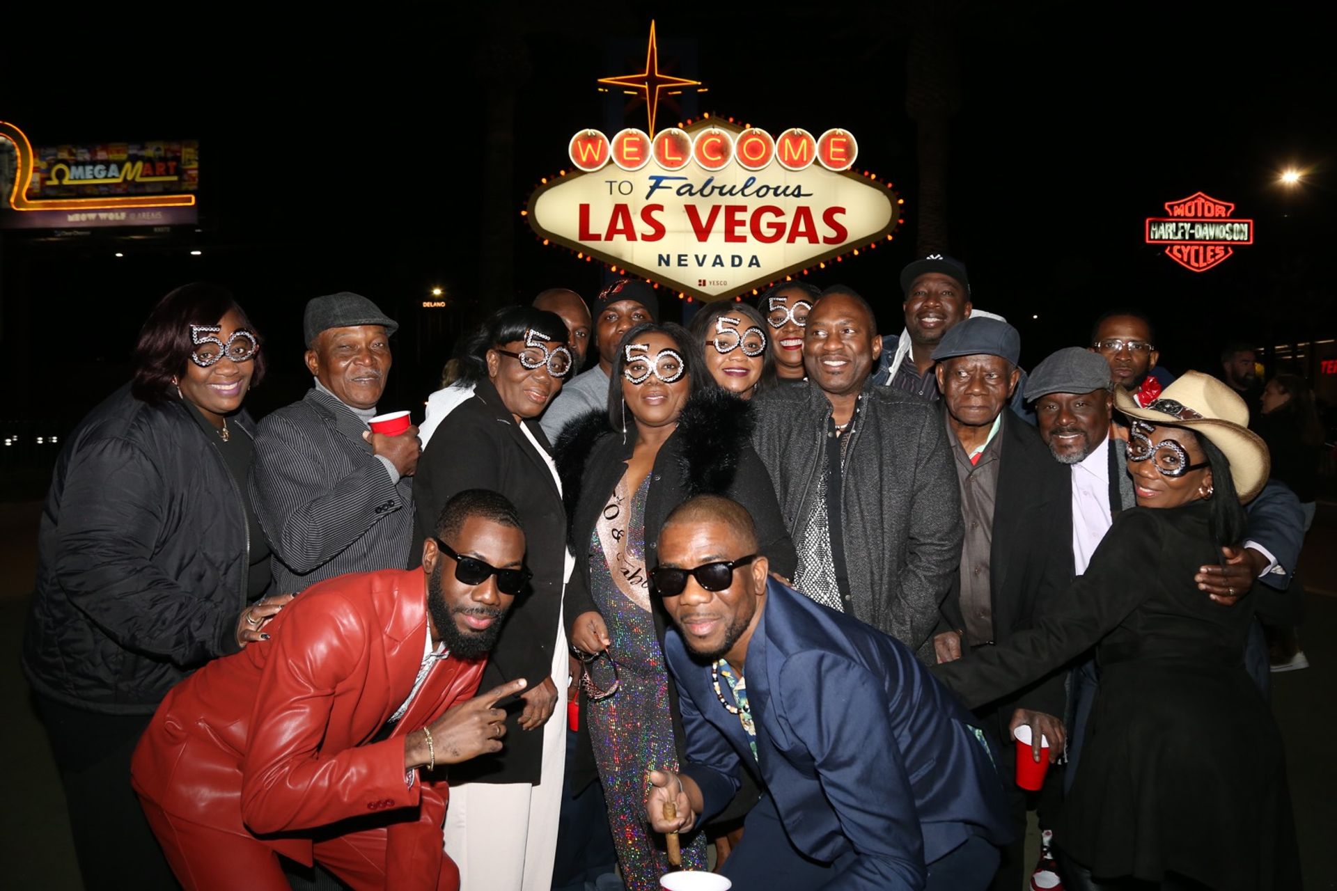Group of people wearing '50' novelty glasses posing at night in front of the Welcome to Fabulous Las Vegas, Nevada sign — festive celebration photo with party outfits and red cups