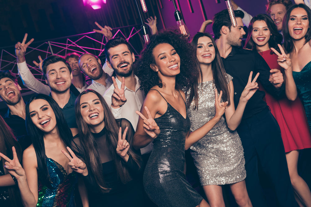Energetic group of young adults at a lively nightclub party, smiling and flashing peace signs under pink neon lights, many wearing sequined dresses and stylish outfits.