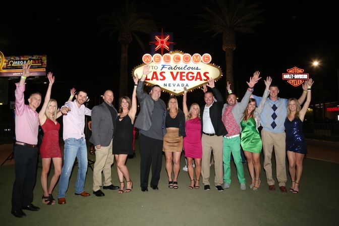 Group of friends cheering with arms raised in front of the iconic 'Welcome to Fabulous Las Vegas Nevada' sign at night, with palm trees and neon lights in the background.