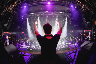 DJ raising arms on stage at a packed nightclub, purple neon lights and smoke beams over turntables and a dancing crowd