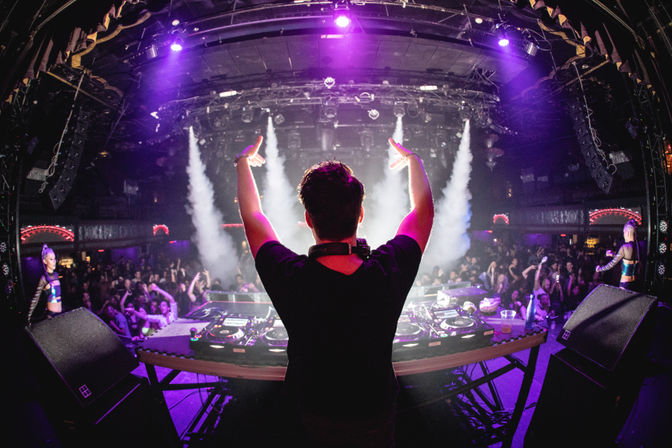 DJ raising arms on stage at a packed nightclub, purple neon lights and smoke beams over turntables and a dancing crowd