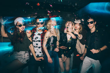 Six young adults posing and dancing at an indoor nightclub party, wearing sunglasses and playful hats, surrounded by falling confetti and colorful neon light streaks.