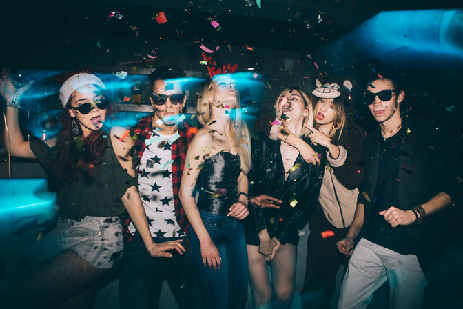 Six young adults posing and dancing at an indoor nightclub party, wearing sunglasses and playful hats, surrounded by falling confetti and colorful neon light streaks.