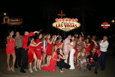 Large celebratory group in red dresses and formalwear at night beneath the Welcome to Fabulous Las Vegas Nevada sign, with a couple kissing at center while friends cheer, pose, and take photos