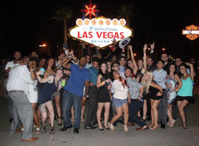 Large group of people partying at night in front of the Welcome to Fabulous Las Vegas, Nevada sign, posing, cheering and holding drinks.