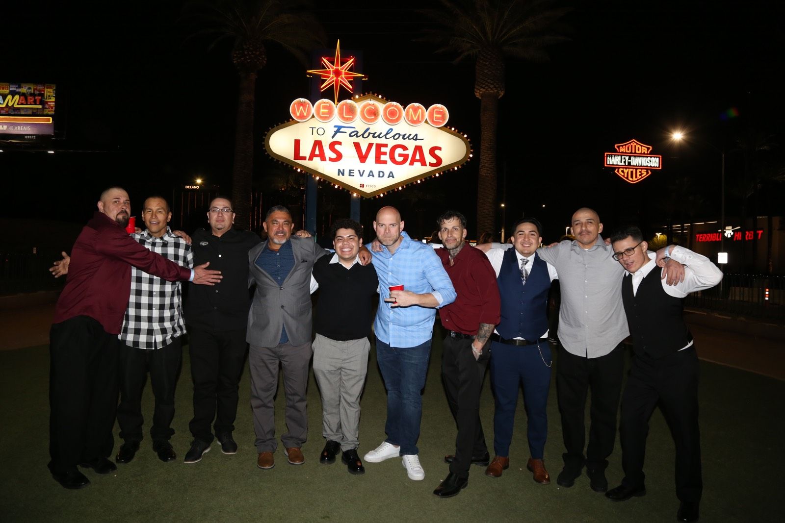 Group of men smiling and posing at night beneath the iconic Welcome to Fabulous Las Vegas Nevada sign, with palm trees and neon lights