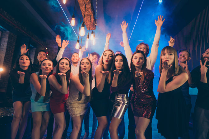 Group of young adults on a nightclub dance floor under hanging Edison bulbs and blue stage lights, women in party dresses blowing kisses while friends cheer.