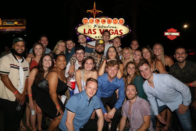 Large group of friends smiling and posing at night in front of the iconic Las Vegas welcome sign, holding drinks and enjoying the nightlife.