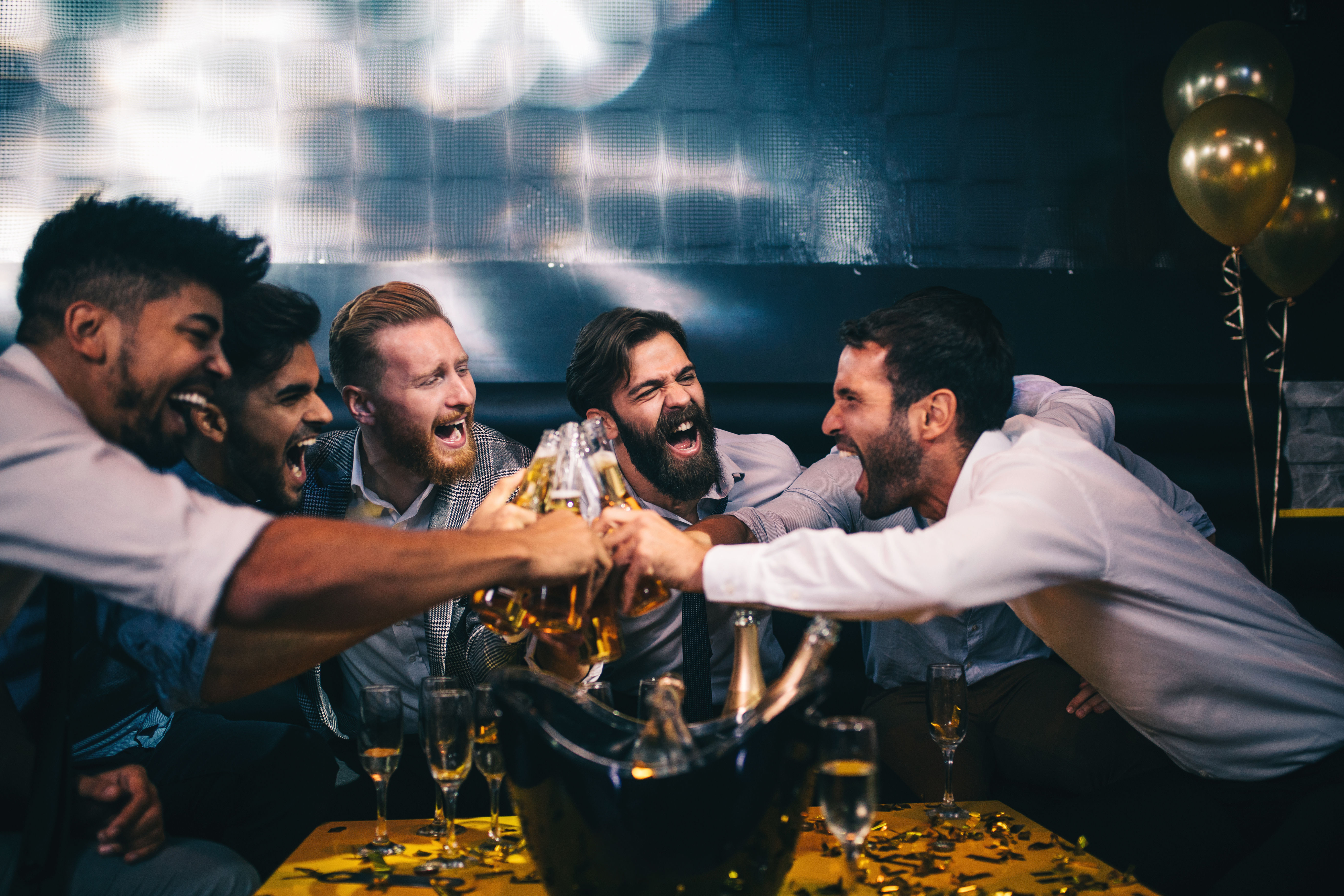 Group of friends cheering and clinking beer bottles in a lively bar lounge, champagne in an ice bucket, glasses, gold confetti and balloons on the table