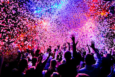 Energetic crowd with hands raised under vivid pink, purple, and blue stage lights as confetti rains down in an indoor live music venue/nightclub.