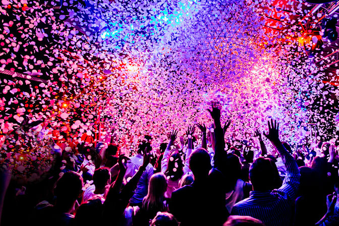 Energetic crowd with hands raised under vivid pink, purple, and blue stage lights as confetti rains down in an indoor live music venue/nightclub.
