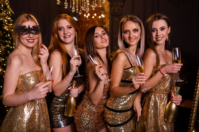 Five women in gold sequined dresses toasting with champagne, one wearing a black masquerade mask, at a glamorous holiday party in an elegant venue