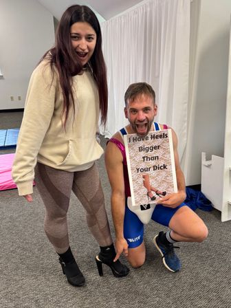 Two adults in an indoor studio: a woman in leggings and black high heels stands while a kneeling man points at her shoe and holds a playful sign reading 'I Have Heels Bigger Than Your Dick'.