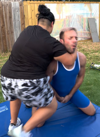 Backyard wrestling scene on a blue mat: person in black shirt and patterned shorts applies a neck hold to a competitor in a blue singlet beside a wooden fence and lawn.