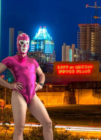 Person in a hot-pink lucha libre mask and matching shiny bodysuit poses confidently at night in front of an illuminated power-plant building and downtown skyscrapers, neon sign glowing and traffic light trails in the foreground.