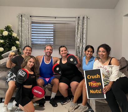 Six friends posing in a bright living room for a home workout photo, sitting on a couch with tiled floor and window blinds, holding boxing focus mitts and a sign that reads 'Crush Them Nuts!,' all smiling and wearing casual workout clothes.