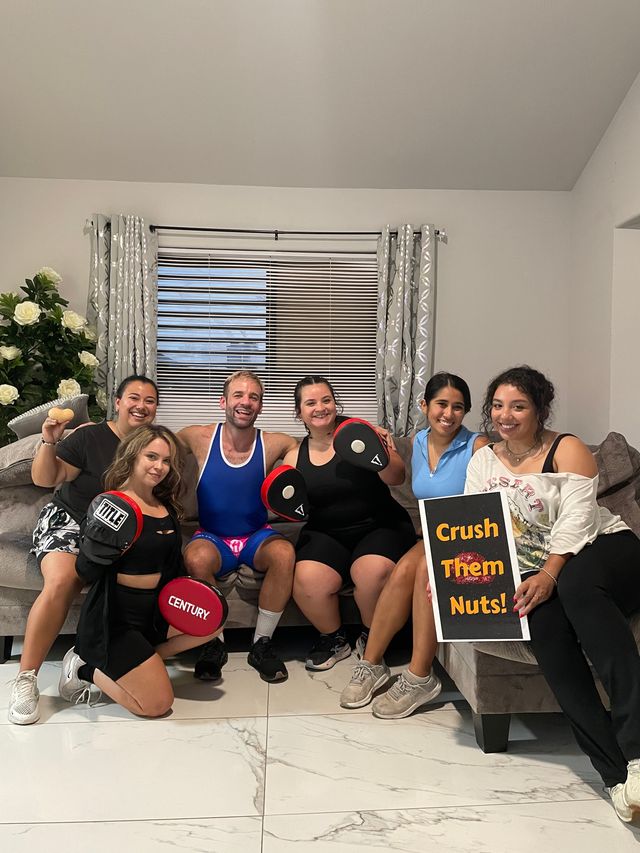 Six friends posing in a bright living room for a home workout photo, sitting on a couch with tiled floor and window blinds, holding boxing focus mitts and a sign that reads 'Crush Them Nuts!,' all smiling and wearing casual workout clothes.