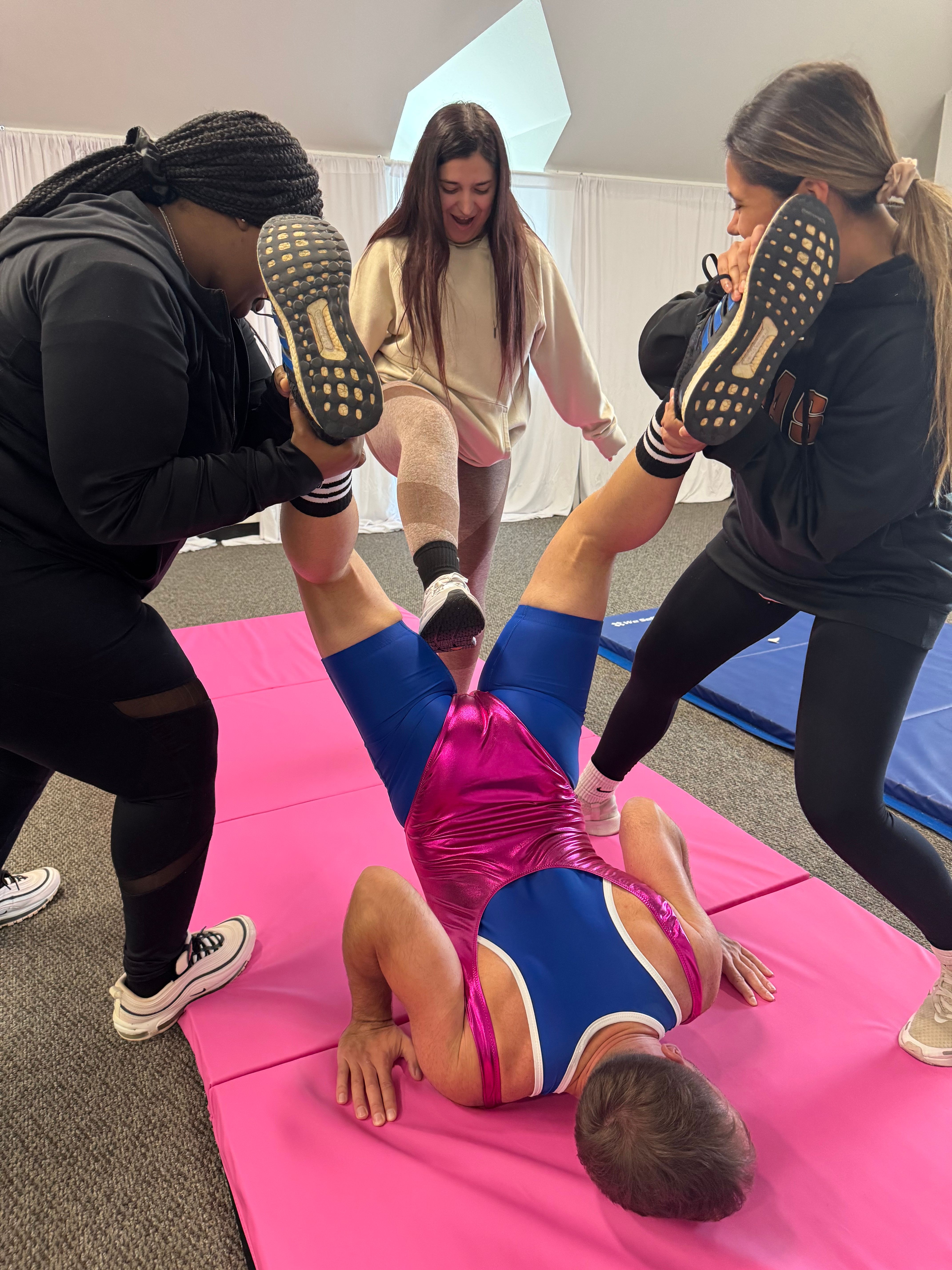 Playful group workout in an indoor fitness studio: three people hold the legs of a person in a shiny pink-and-blue singlet doing a bridge on a pink mat.