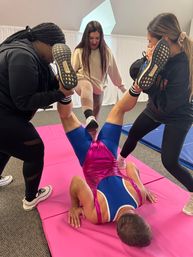 Playful group workout in an indoor fitness studio: three people hold the legs of a person in a shiny pink-and-blue singlet doing a bridge on a pink mat.