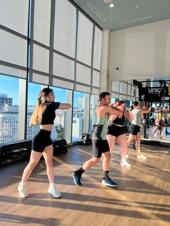 Energetic sunlit high-rise fitness studio with four people in a group cardio/boxing workout, punching in sync by floor-to-ceiling windows overlooking a downtown city skyline.
