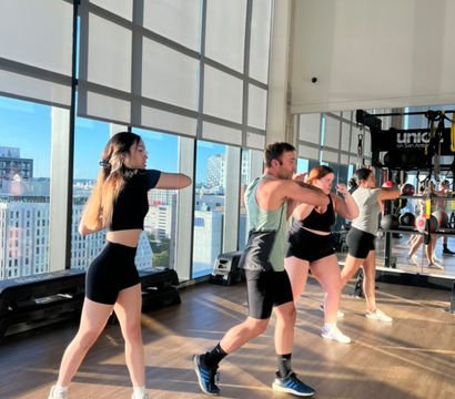 Energetic group fitness class practicing punches in a high-rise gym studio with floor-to-ceiling windows, urban skyline view, mirrored wall, and sunlit hardwood floor.