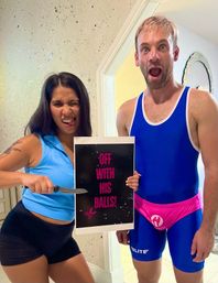 Playful couple in a home hallway: woman in a blue tank and black shorts mock-wields a knife at a sign that reads 'OFF WITH HIS BALLS!', while a surprised man in a blue singlet and bright pink athletic supporter reacts.