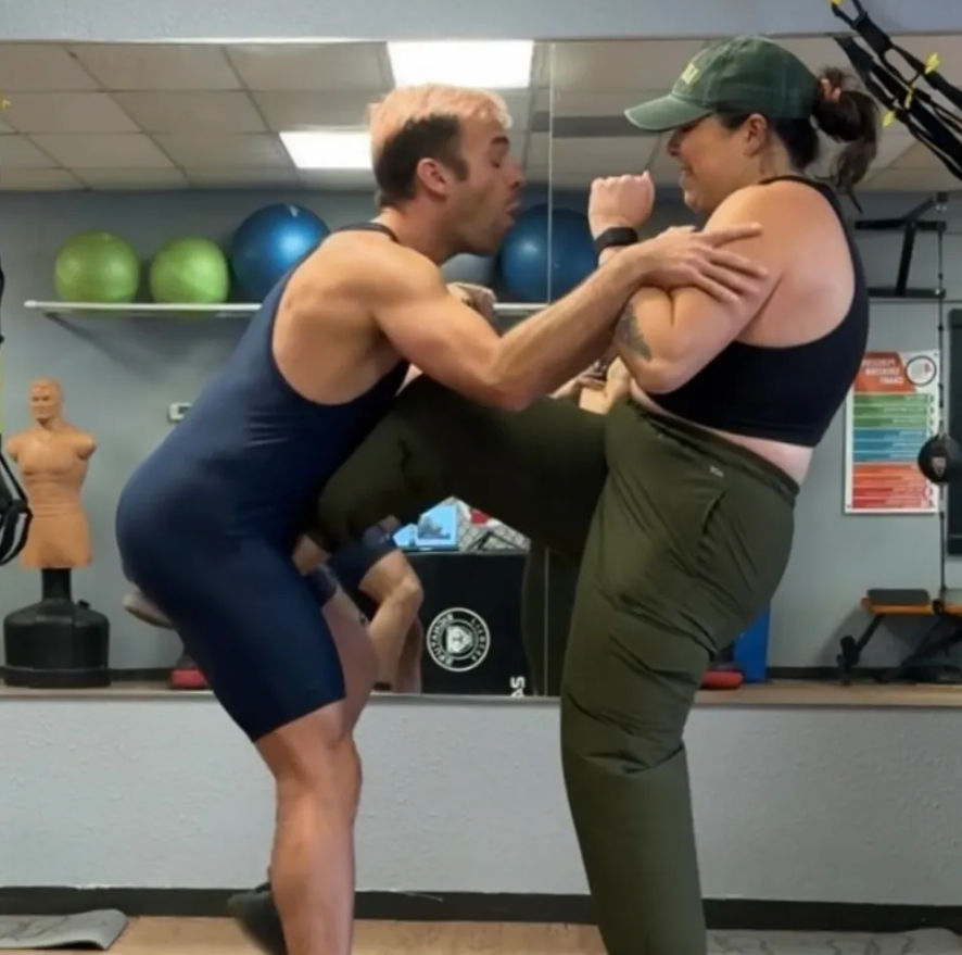 Woman delivers a knee strike to a partner during self-defense training in a mirrored gym studio with exercise balls and a training dummy.