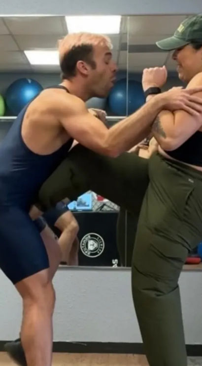 Woman delivers a knee strike to a partner during self-defense training in a mirrored gym studio with exercise balls and a training dummy.