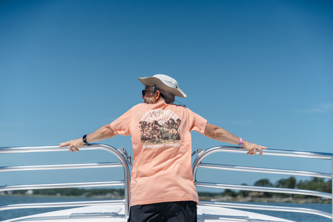 Person wearing a sun hat and peach graphic T-shirt leans on a boat railing, enjoying a sunny boat ride near a tree-lined shoreline under a clear blue sky.