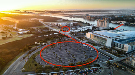 Aerial sunrise view of a riverfront convention center and marina with a large empty parking lot circled “Park here” and a marked “Meet here” near a ferry