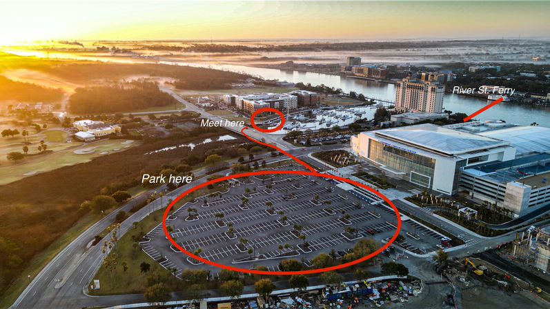 Aerial sunrise view of a riverfront convention center and marina with a large empty parking lot circled “Park here” and a marked “Meet here” near a ferry