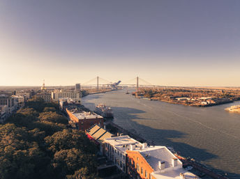 Aerial golden-hour view of the Savannah riverfront: sunlit historic brick buildings and oak-lined waterfront with a docked riverboat and the cable-stayed Talmadge Memorial Bridge spanning the river under a clear sky.