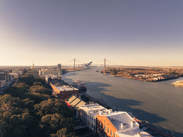 Aerial golden-hour view of the Savannah riverfront: sunlit historic brick buildings and oak-lined waterfront with a docked riverboat and the cable-stayed Talmadge Memorial Bridge spanning the river under a clear sky.