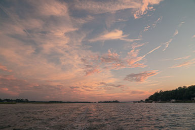 Pink and orange sunset clouds over a coastal estuary, rippling water with a boat wake in the foreground and tree-lined shoreline with docks on the right